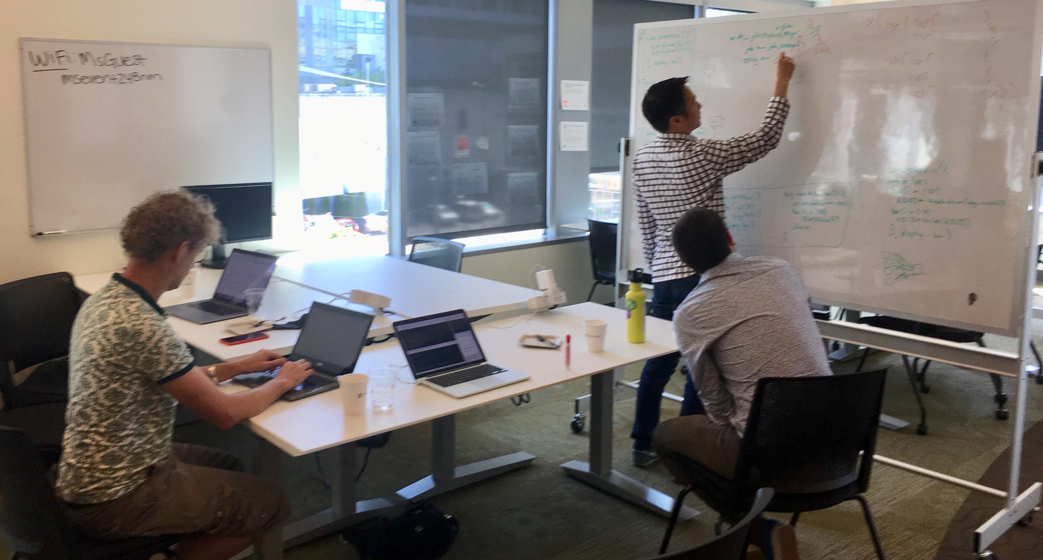 One person writing on a whiteboard, two people at a table